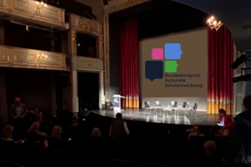 2025_06_News_Bundeskongress_Bamberg Saal des Bamberger ETA-Hoffmann-Theaters, eine beleuchtete Bühne mit einer Leinwand zeigt das Logo des Bundeskongress kulturelle Schulentwicklung zu sehen, die