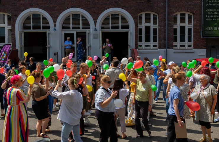 Eine große Personengruppe geht mit bunten Luftballons kreuz und quer über einen Platz
